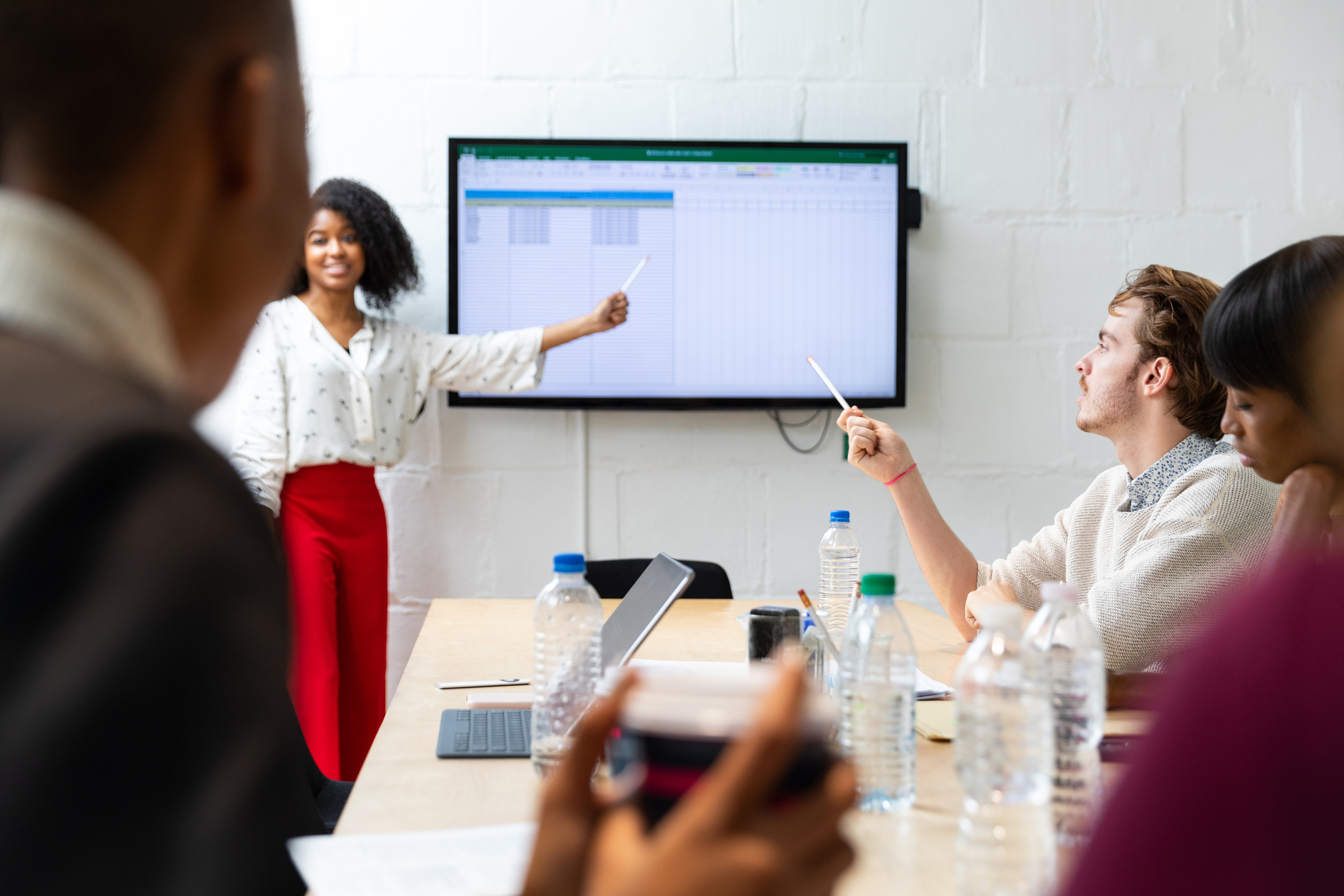 5 Reasons Excel  Could Be Holding Your Business Back (a woman presents to her team in a conference room, pointing to an Excel spreadsheet on a large screen on the wall)